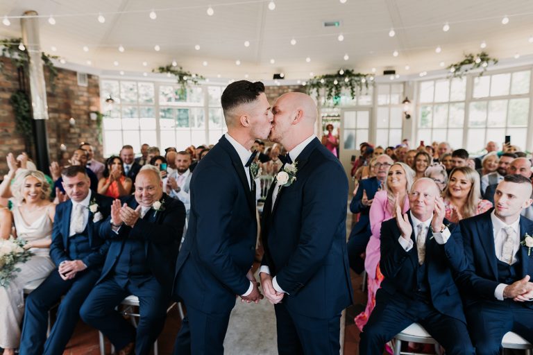 Gay wedding at Galgorm Resort hotel and spa in Ballymena. Both grooms kiss and hold hands they are now husband and husband. Showing all guests in background.
