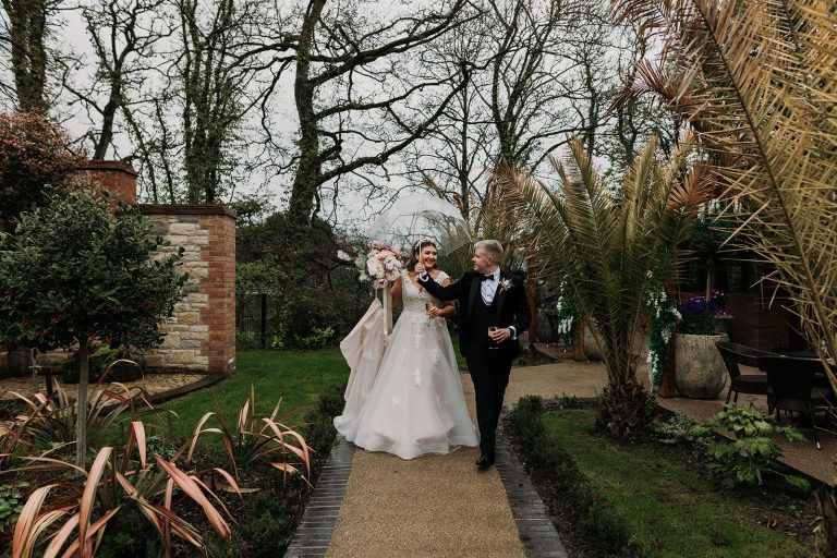 Bride and groom walking in rain with clear umbrella's in The Loft wedding venue gardens