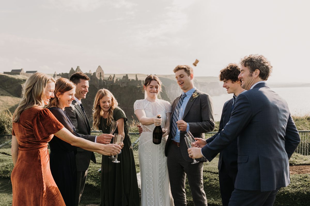 eloping couple standing on cliff top with Dunluce castle behind them. they have a few members of their family around as they pop open a bottle of champagne the cork pops and visible in photo
