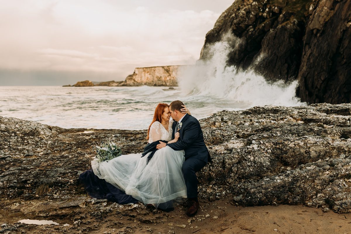 bride and groom at Ballintoy sitting on rocks with big waves behind them
