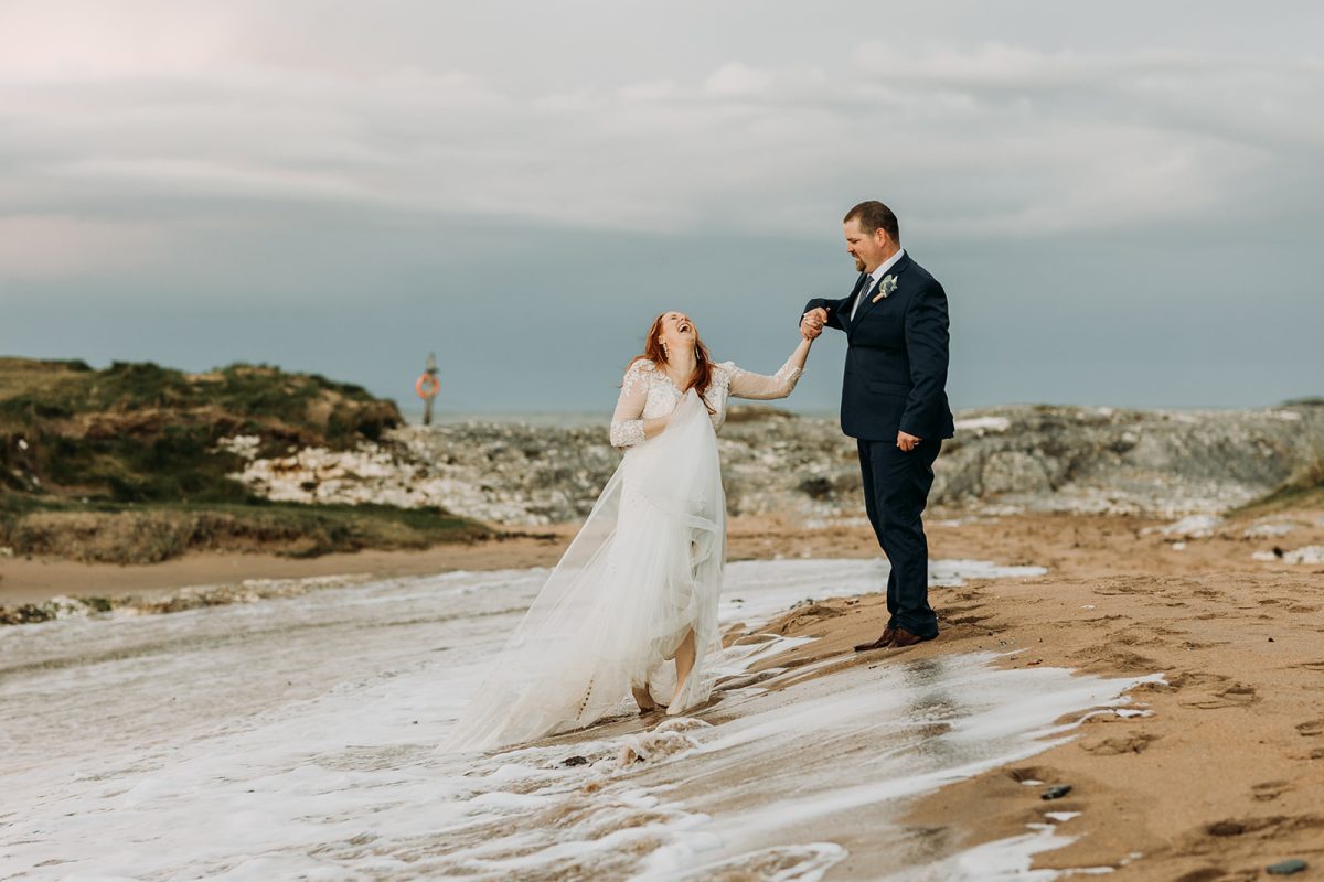 beide and groom walking on the beach and the wave has came in and wet their feet. bride laughing