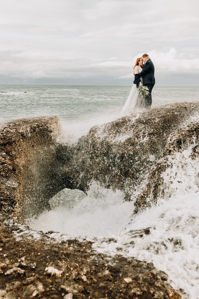 bride and groom standing on cliff with sea backdrop. in the rock is a massive hole and t4h wave has came up through hole and soaked both bride and groom