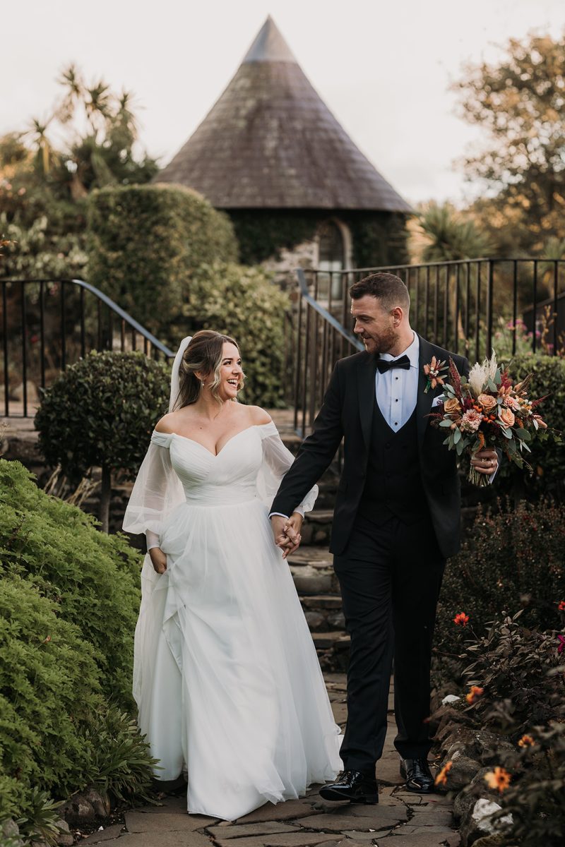 Bride and groom walking along pathway surrounded by flowers at Ballygally Castle