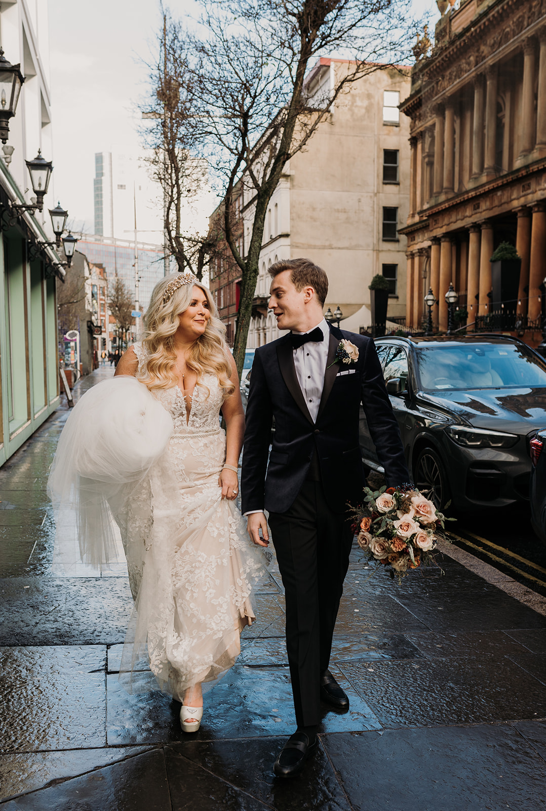 bride holding her dress up as she and groom walks in belfast streets. The Merchant hotel is in background.