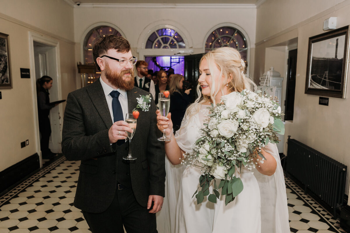 bride and groom cheers their fizz glass together with strawberries on top as they walk out of their ceremony room just married.