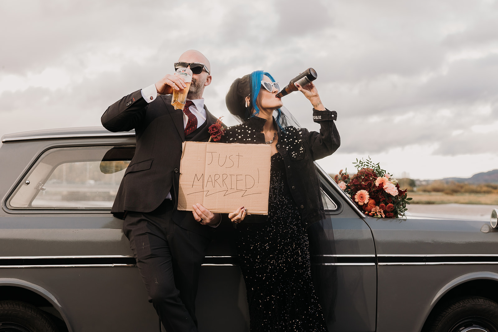 bride and groom on wedding day. bride wears black dress. both are leaning against a vintage car drink beer holding a sign saying, "just married".
